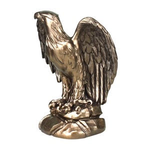 Pode incluir: Uma estatueta de &aacute;guia na cor bronze sobre uma base de rocha. A &aacute;guia &eacute; representada com a cabe&ccedil;a virada para o lado e as asas parcialmente abertas, exibindo texturas detalhadas de penas e um acabamento met&aacute;lico em toda a escultura.