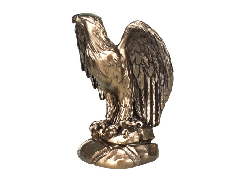Pode incluir: Uma estatueta de &aacute;guia na cor bronze sobre uma base de rocha. A &aacute;guia &eacute; representada com a cabe&ccedil;a virada para o lado e as asas parcialmente abertas, exibindo texturas detalhadas de penas e um acabamento met&aacute;lico em toda a escultura.
