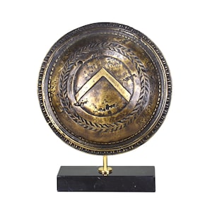 Puede incluir: Réplica de escudo espartano de color bronce, con un diseño de galón dorado y corona de laurel. El escudo está montado sobre una base de mármol negro con un soporte dorado, creando una pieza decorativa histórica.