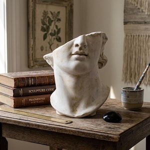 May include: A weathered, off-white bust of a classical figure, missing a portion of the head, sits on a rustic wooden table. Beside it are stacked antique books and a small ceramic cup with a paintbrush. A black stone and a brass ruler are also on the table.