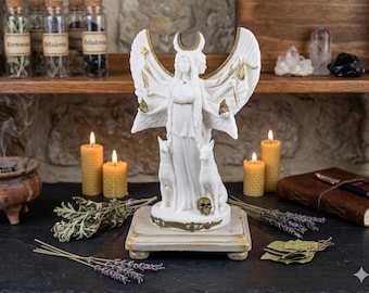 May include: A white statue of a goddess with outstretched wings, a crescent moon headdress, and two cat figures. The statue sits on a decorative base with gold accents. Lit candles, herbs, and a leather-bound journal surround the statue.