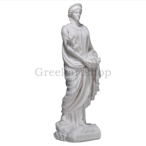 May include: A white marble statue of Demeter, the Greek goddess of agriculture. She is depicted in flowing robes, holding a bundle of wheat and a basket of flowers. The statue is on a stone base, with the name "DEMETER" inscribed.