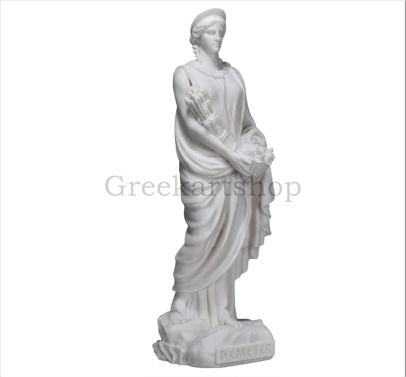 May include: A white marble statue of Demeter, the Greek goddess of agriculture. She is depicted in flowing robes, holding a bundle of wheat and a basket of flowers. The statue is on a stone base, with the name "DEMETER" inscribed.