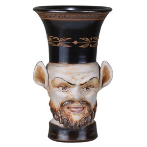 Puede incluir: Un jarrón de cerámica con la parte superior negra y un diseño de rostro. La cara tiene barba, orejas grandes y una expresión enfadada. El jarrón tiene una base marrón oscuro y está decorado con detalles dorados.