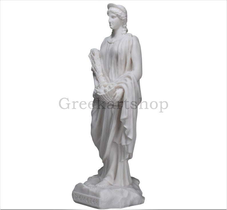 May include: A white marble statue of Demeter, the Greek goddess of agriculture. She is depicted in flowing robes, holding a basket of fruits and wheat. The statue has the text "Demeter" at the base and the text "Greekartshop" in the background.