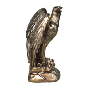 Pode incluir: Uma est&aacute;tua de &aacute;guia na cor bronze sobre uma base rochosa. A ave &eacute; representada de perfil com as asas parcialmente dobradas contra o corpo, exibindo texturas detalhadas das penas e um olhar atento direcionado para o lado direito.