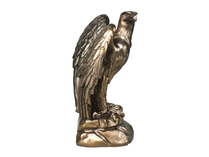 Pode incluir: Uma est&aacute;tua de &aacute;guia na cor bronze sobre uma base rochosa. A ave &eacute; representada de perfil com as asas parcialmente dobradas contra o corpo, exibindo texturas detalhadas das penas e um olhar atento direcionado para o lado direito.