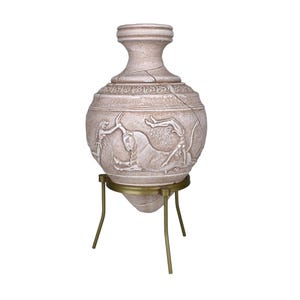Könnte beinhalten: Eine beige Vase im antiken Stil mit einem detaillierten Relief von Figuren und einem Stier, montiert auf einem messingfarbenen Dreibeinständer. Die Vase hat einen rissigen Anschein, was zu ihrer gealterten Ästhetik beiträgt. Das Design erinnert an antike griechische Keramik.