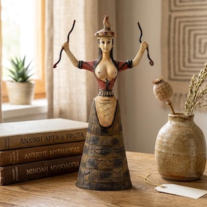 May include: A decorative figurine of a woman with arms raised, holding curved objects. The figure wears a red and brown top and a patterned skirt. A small bird sits atop her head. Books and a vase with dried plants are in the background.