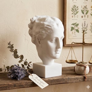 Peut inclure: Buste en plâtre blanc d'une tête de femme sur une base carrée. Le buste est posé sur une étagère en bois avec une branche de lavande et une étiquette indiquant "Handcrafted Goddess of Health". Une balance vintage et une impression botanique sont en arrière-plan.