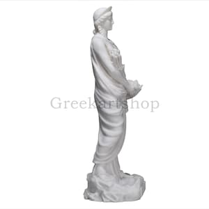 May include: A white marble statue of a woman in classical attire, holding flowers in a basket. The statue has a serene expression and is set against a white background. The text "Greekartshop" is visible.