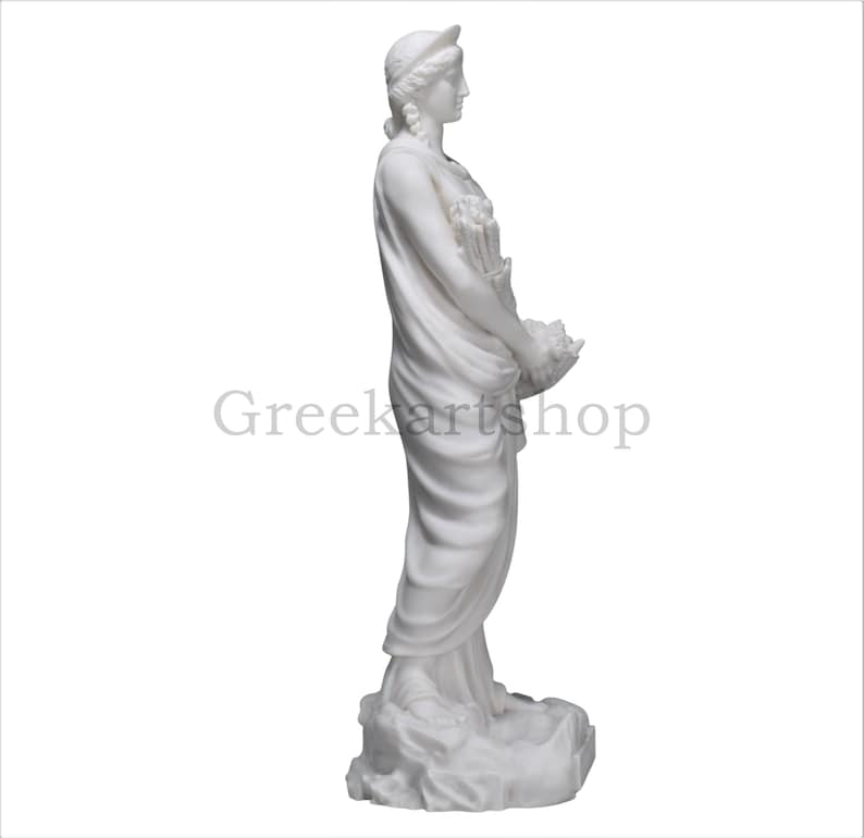 May include: A white marble statue of a woman in classical attire, holding flowers in a basket. The statue has a serene expression and is set against a white background. The text "Greekartshop" is visible.