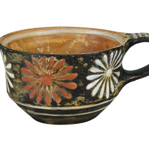 Puede incluir: Una taza de té de cerámica con asa, con un exterior negro adornado con diseños florales blancos y naranjas. El interior tiene un tono cálido y terroso. La taza tiene una base redondeada y una abertura ancha.