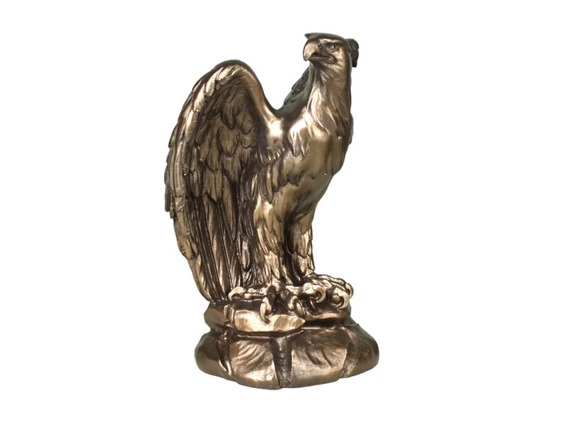 Pode incluir: Uma estatueta de &aacute;guia na cor bronze sobre uma base de rocha texturizada. A &aacute;guia &eacute; representada de perfil com as asas parcialmente abertas, exibindo texturas detalhadas nas penas e um bico afiado, com um acabamento de brilho met&aacute;lico.
