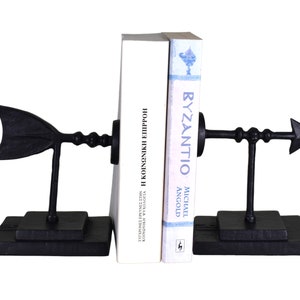 May include: A pair of black metal bookends shaped like arrows. The bookends are holding up two books, one with the title "Byzantio" and the other with the title "Η ΚΟΙΝΩΝΙΚΗ ΕΠΙΡΡΟΗ".