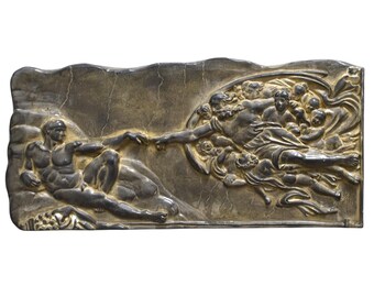 May include: A rectangular, metallic relief sculpture depicting Michelangelo's "The Creation of Adam." The artwork features two figures with outstretched arms, nearly touching. The relief is in shades of gray and gold, with detailed figures and a textured background.