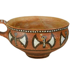 Puede incluir: Una taza de cerámica de color terracota con asa. La taza está decorada con patrones geométricos blancos y negros, incluyendo formas de hacha estilizadas y líneas punteadas. La taza tiene una abertura ancha y un pie pequeño.