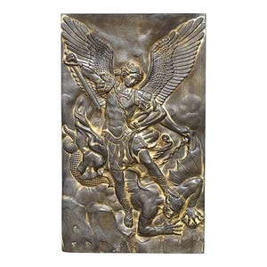 May include: A rectangular wall plaque featuring a metallic bronze-toned relief sculpture of an angel with large wings, wearing armour and holding a sword, standing over a winged creature. The design has dark recessed areas and highlighted edges.