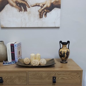 Puede incluir: Una c&oacute;moda de madera con dos cajones y patas de metal negro. La c&oacute;moda tiene un acabado de madera de grano marr&oacute;n claro. Un jarr&oacute;n de cer&aacute;mica negro y marr&oacute;n con una manija negra se encuentra sobre la c&oacute;moda. El jarr&oacute;n tiene un dise&ntilde;o en blanco y negro de figuras sobre un caballo. Tres bolas de mimbre tejidas y dos velas blancas se encuentran sobre una bandeja de metal dorado frente al jarr&oacute;n. Una impresi&oacute;n enmarcada de una pintura de dos manos que se alcanzan entre s&iacute; cuelga en la pared detr&aacute;s de la c&oacute;moda.