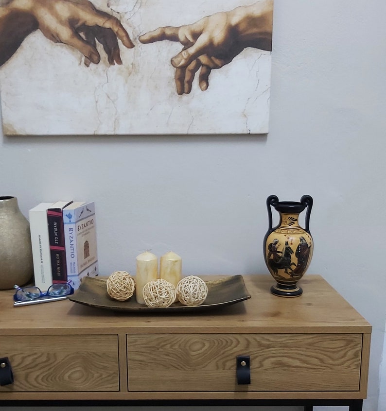 Puede incluir: Una c&oacute;moda de madera con dos cajones y patas de metal negro. La c&oacute;moda tiene un acabado de madera de grano marr&oacute;n claro. Un jarr&oacute;n de cer&aacute;mica negro y marr&oacute;n con una manija negra se encuentra sobre la c&oacute;moda. El jarr&oacute;n tiene un dise&ntilde;o en blanco y negro de figuras sobre un caballo. Tres bolas de mimbre tejidas y dos velas blancas se encuentran sobre una bandeja de metal dorado frente al jarr&oacute;n. Una impresi&oacute;n enmarcada de una pintura de dos manos que se alcanzan entre s&iacute; cuelga en la pared detr&aacute;s de la c&oacute;moda.