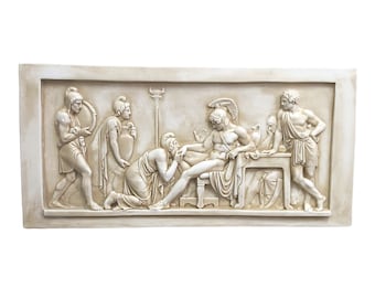 May include: A white plaster relief sculpture depicting a scene from Greek mythology. The sculpture shows a group of figures, including a man lying on a couch, a woman kneeling before him, and several other figures standing around them.