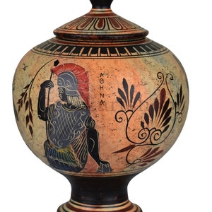 May include: A ceramic Greek urn with a lid, decorated with a black-figure painting of a warrior holding a spear. The urn is painted in shades of red, black, white, and yellow. The urn has a round body and a tall, slender base. The urn is decorated with a floral pattern and the word "ATHENA" in Greek.
