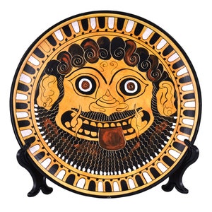 Może przedstawiać: Ceramiczny talerz z czarno-białym obramowaniem i namalowanym obrazem twarzy Gorgony w środku. Twarz Gorgony jest żółta z czarnymi włosami, dużym językiem i dużymi oczami.