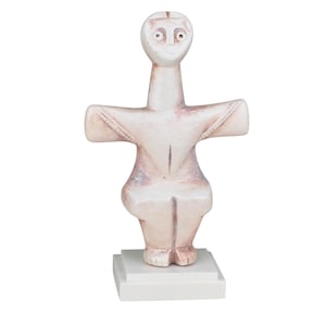 May include: A light-colored, stylized sculpture of a figure with outstretched arms, mounted on a white square base. The sculpture has a simple, abstract design with a smooth texture and subtle pinkish hues.