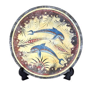 Pode incluir: Prato decorativo redondo com dois golfinhos azuis nadando em um fundo amarelo. O prato tem uma borda marrom escura com um padrão decorativo e é exibido em um suporte preto. Os golfinhos são detalhados com detalhes azuis e brancos.