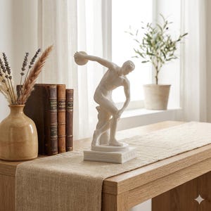 May include: A white matte finish sculpture of a discus thrower standing on a square base with a Greek key pattern. The figure is depicted in a dynamic athletic pose, captured mid-motion with one arm extended holding a disc and the other bent.