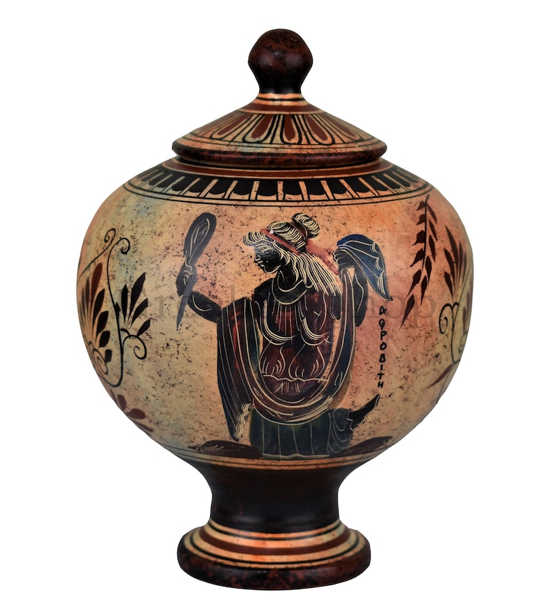 May include: A brown ceramic Greek style urn with a lid. The urn is decorated with a black and white image of a woman holding a mirror. The text "APHRODITE" is written on the urn.