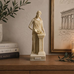 Może przedstawiać: Kremowa statua klasycznej postaci kobiecej trzymającej misę, stojącej na cokole z napisem "ESTIA". Statua stoi na drewnianej powierzchni, z oprawionym wydrukiem architektonicznym i zapaloną świecą w tle.