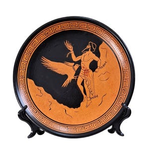 Op de afbeelding: Zwart en oranje decoratief bord met een Griekse mythologische scène. Het bord heeft een figuur met een adelaar en een geometrische rand. Het bord wordt gepresenteerd op een zwarte standaard.