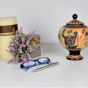 May include: A decorative ceramic urn with a lid, painted with a scene of a woman holding a mirror and a shield. The urn is brown and beige with black and blue accents. The urn is on a white surface with a pair of blue glasses and a silver pen.