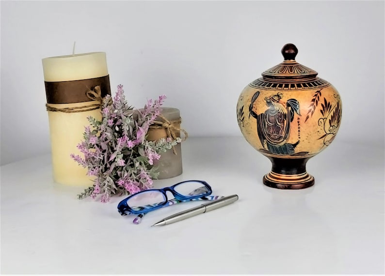 May include: A decorative ceramic urn with a lid, painted with a scene of a woman holding a mirror and a shield. The urn is brown and beige with black and blue accents. The urn is on a white surface with a pair of blue glasses and a silver pen.