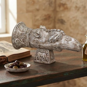 Può includere: Una scultura in resina color argento dall'aspetto invecchiato raffigurante una testa di grifone montata su una base rettangolare con un rilievo di cervo. La scultura presenta volute complesse sul collo e una finitura testurizzata, posata in orizzontale.