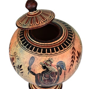 May include: A ceramic Greek-style urn with a lid, decorated with black and red geometric patterns and a painted image of a woman holding a mirror. The urn is on a white background.
