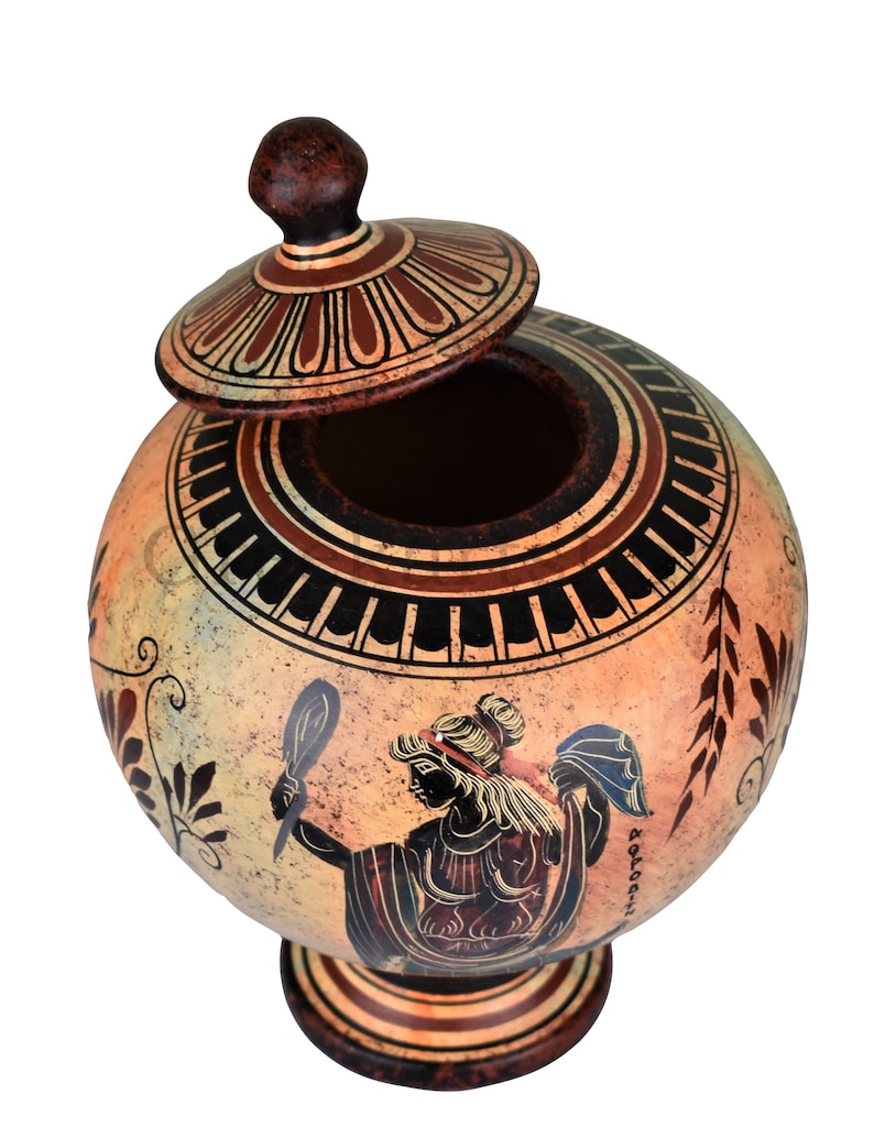 May include: A ceramic Greek-style urn with a lid, decorated with black and red geometric patterns and a painted image of a woman holding a mirror. The urn is on a white background.