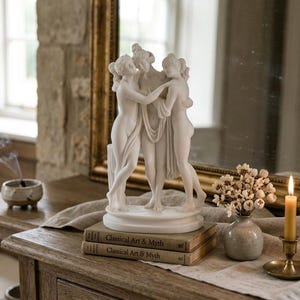 Puede incluir: Una escultura de m&aacute;rmol blanco de tres figuras abraz&aacute;ndose, colocada sobre una mesa de madera con libros, un jarr&oacute;n con flores y una vela encendida. La escultura es el punto focal, con un espejo en el fondo.
