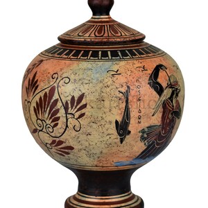 May include: A ceramic Greek amphora with a lid, decorated with a red and black floral design, a fish, and a figure with a staff. The amphora is on a white background.