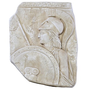 Op de afbeelding: Een beige steenkleurige wandplaat met een reliëf van een figuur met een helm met kam, die een speer en een rond schild versierd met een uil vasthoudt. De bovenrand toont een Grieks sleutelpatroon en het oppervlak heeft een verweerde textuur.