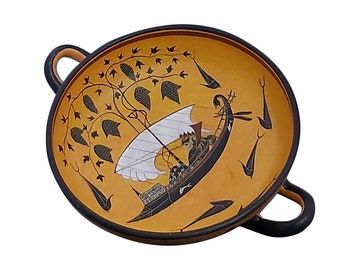 May include: A black and orange ceramic bowl with two handles. The bowl features a painted design of a ship with a white sail and a figure on board, surrounded by stylized leaves and fish.