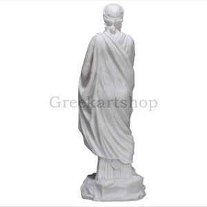 May include: A white marble statue of a draped figure, viewed from the back. The figure wears a head covering and a flowing garment. The statue is on a rocky base. The text "Greekartshop" is visible.