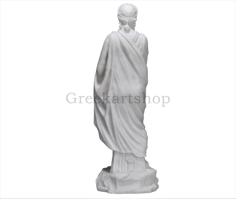 May include: A white marble statue of a draped figure, viewed from the back. The figure wears a head covering and a flowing garment. The statue is on a rocky base. The text "Greekartshop" is visible.
