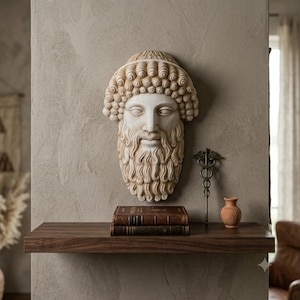 May include: A cream-coloured, sculpted wall hanging of a bearded man's face with curly hair. A bronze medical symbol and a small terracotta vase sit on a wooden shelf below. Two antique books are also on the shelf.