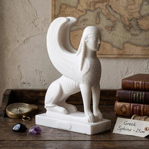 Op de afbeelding: Een witte marmeren sfinx-standbeeld met grote vleugels, zittend op een rechthoekige basis. Het beeld is ongeveer 21 cm hoog en is gelabeld "Greek Sphinx - 21cm". De achtergrond omvat antieke boeken, een kompas en een vintage kaart.