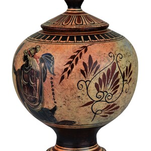 May include: A ceramic Greek style urn with a lid, painted with a figure of a woman holding a staff and a floral design. The urn is decorated with red, black, and white paint on a beige background. The text "&Alpha;&Phi;&Rho;&Omicron;&Delta;&Iota;&Tau;&Eta;" is written on the side of the urn.