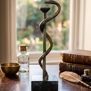 Könnte beinhalten: Eine bronzefarbene Skulptur des Äskulapstabs mit einer um einen zentralen Stab gewundenen Schlange, montiert auf einem quadratischen Sockel aus dunkelgrünem Marmor. Die Metallfigur besitzt eine kleine kreisförmige Plattform an der Spitze des Stabes.