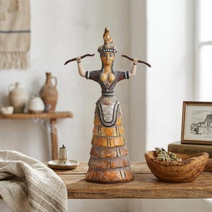 May include: A decorative figurine of a Minoan snake goddess with a tall, tiered yellow and brown skirt, a dark bodice, and a headdress. The figure holds two dark, curved snake-like objects in outstretched arms, featuring painted facial details.