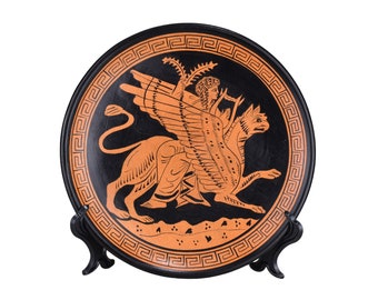 Może przedstawiać: Czarny i pomarańczowy ceramiczny talerz z greckim kluczem na krawędzi. Talerz przedstawia wizerunek skrzydlatego stworzenia z ludzką głową i lwią sylwetką.